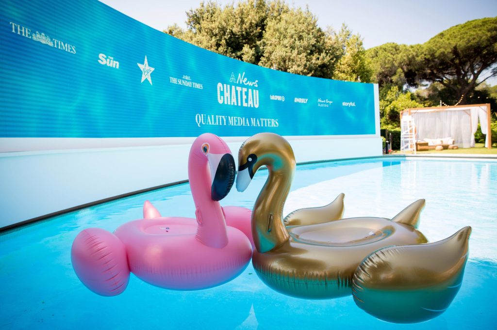 News Chateau curved LCD screen showing brand logos, positioned around pool with inflated flamingo swimming toys as props