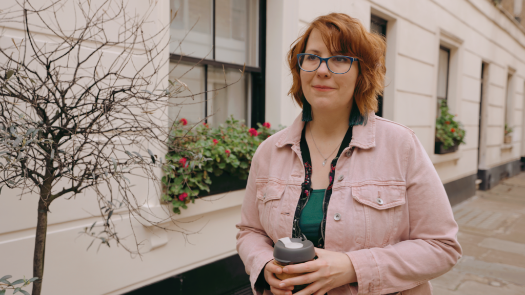 Woman smiles off camera in London lane, wearing pink jacket